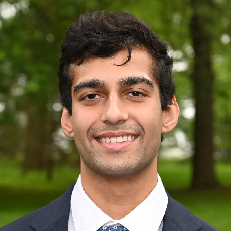 Nikhil Ramlukan, smiling, in a navy suit and white shirt, outdoors in front of trees.