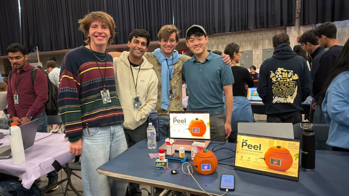 The four of us at the HackMIT judging table, arms round each other, behind the orange instrument, a laptop and a second screen both showing the Peel title card, the phone on the Peel scan screen, and a scatter of tablets and pill bottles on the tablecloth.